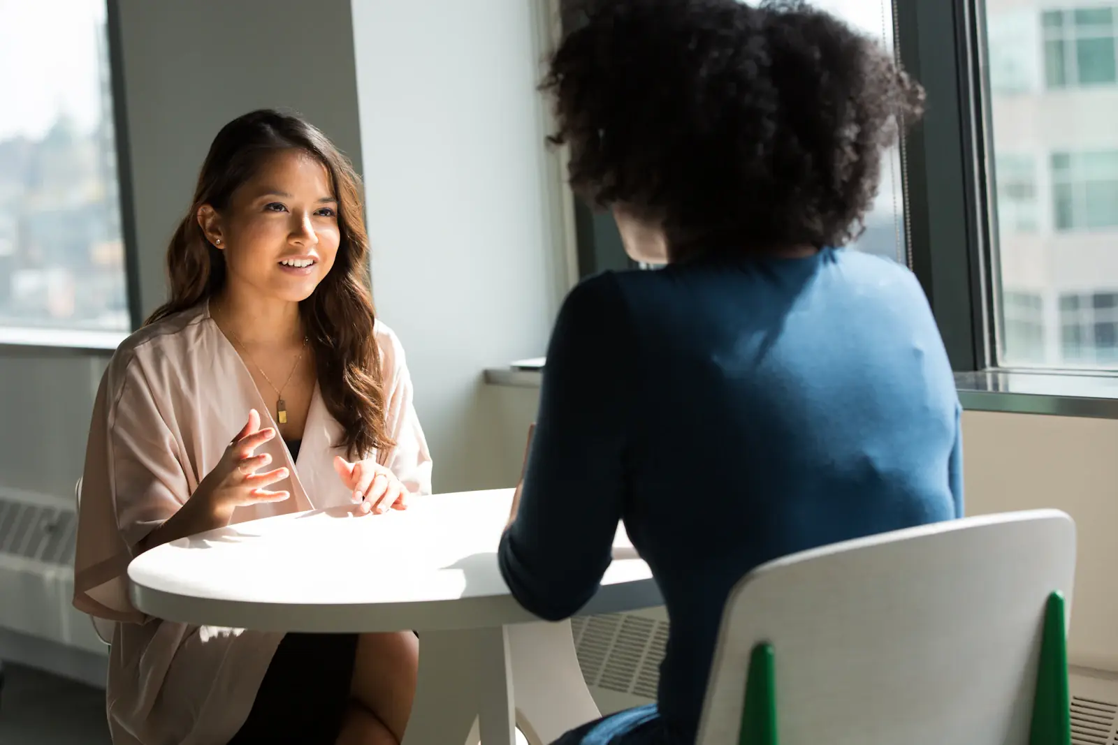 Twee vrouwen in gesprek aan een ronde tafel bij een raam in een licht kantoor
