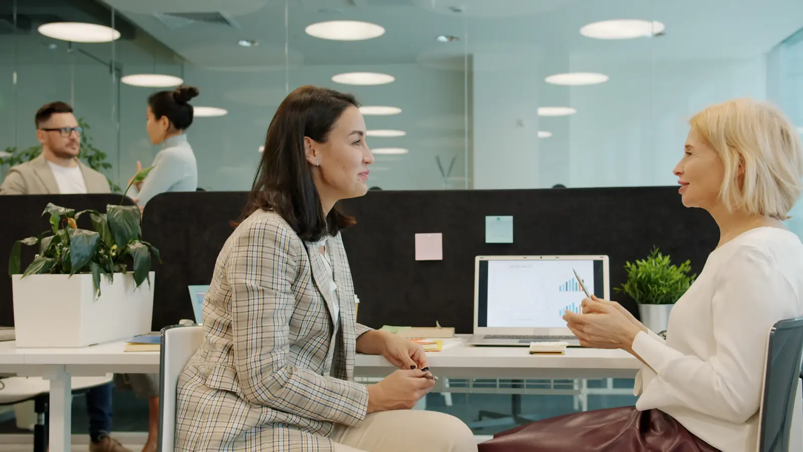 Twee collega’s bespreken iets aan een bureau in een modern kantoor met een laptop tussen hen in